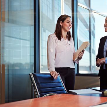 Two professional women smiling and discussing in a modern office room.