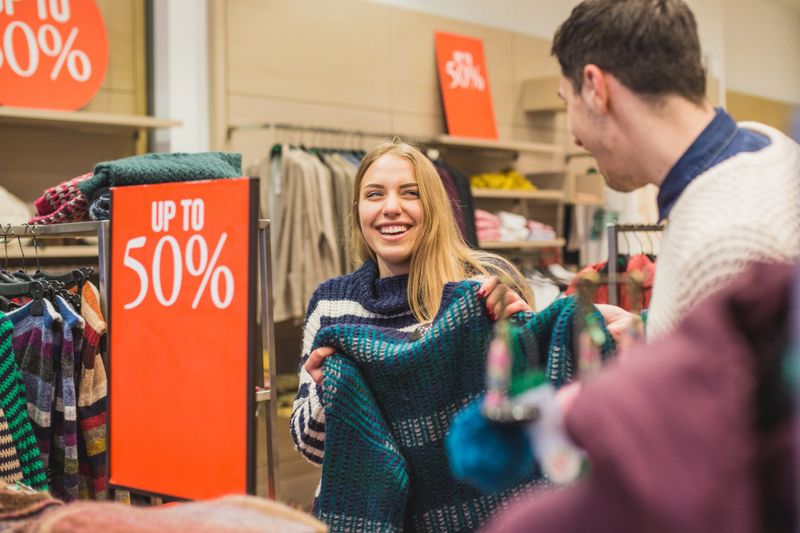 Woman showing new shirt to young man while shopping for clothes, they are standing next to the sale sign in store, girl is laughing.