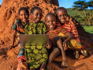 Four children sitting outdoors, smiling with a laptop on their laps.
