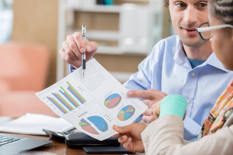Confident Caucasian male accountant meets with African American female associate. He is showing her something on a document containing charts and graphs.