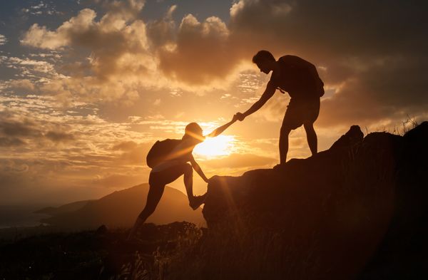 Two hikers helping each other climb a rocky hill at sunset.