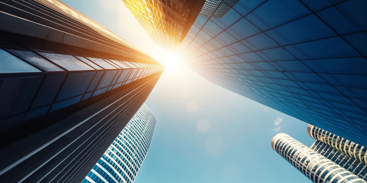 Sunlight shines between tall modern skyscrapers under a clear blue sky.
