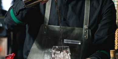 Bartender pouring a dark drink over ice in a glass.