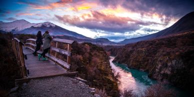 Two people standing on a wooden bridge overlooking a scenic river and mountains.