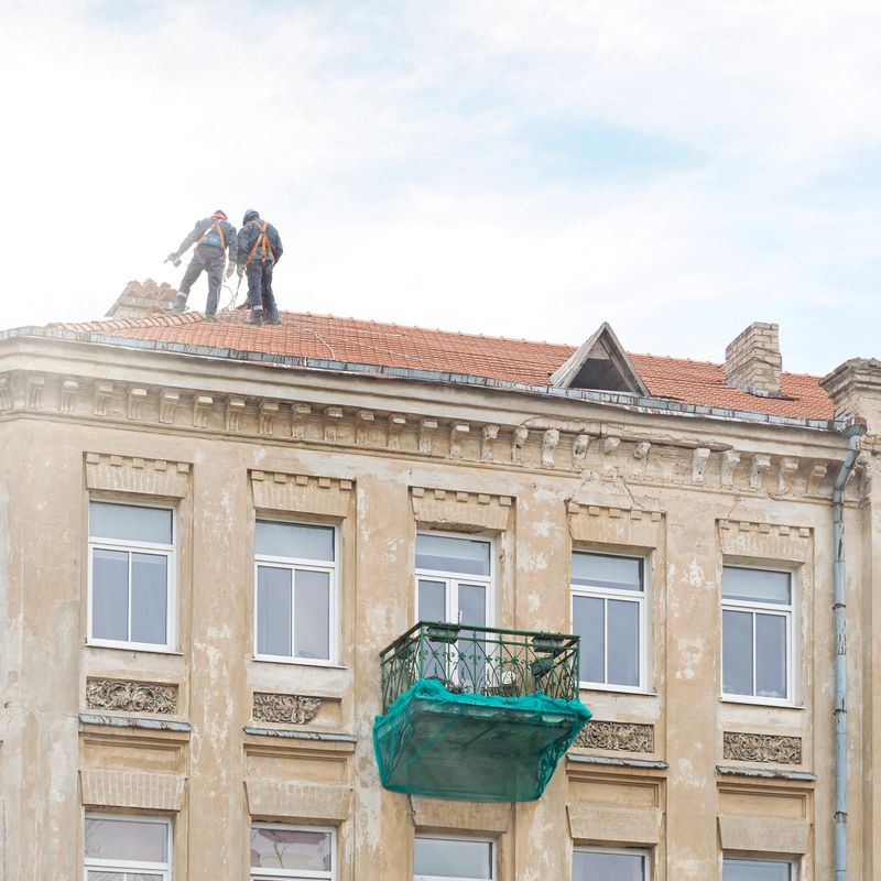 Roof repair of old building, two workers on a roof with electric drill, cracked balcony in green net