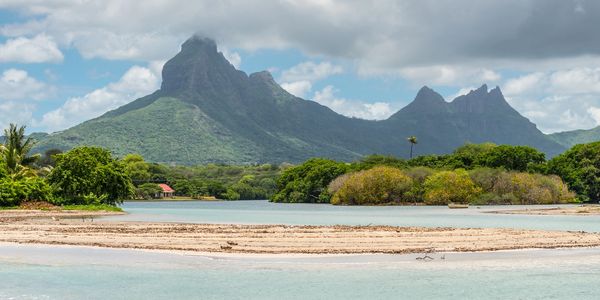 Tropical island scenery with mountains, lush greenery, and calm waters under a cloudy sky.