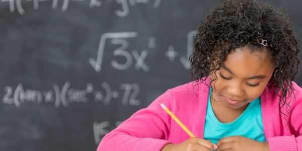 A young girl writing while sitting in front of a chalkboard with math equations.