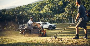 Two men mowing grass on a sunny day in a tropical setting.