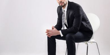 Man in black suit sitting on a white chair against a plain background.