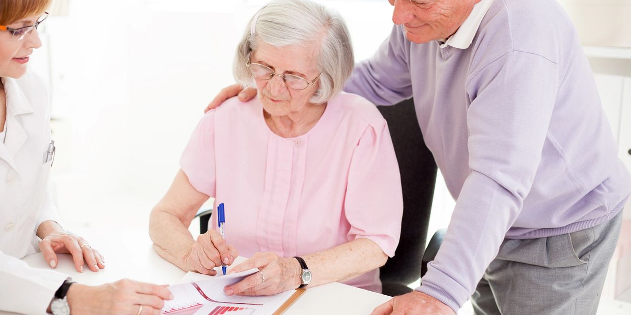 Elderly woman signing documents with support from a man and a professional.