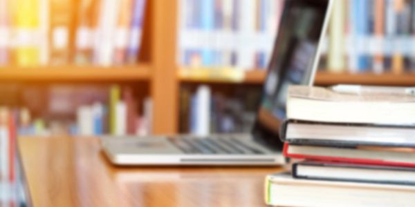 Laptop and stack of books on a wooden table in a library.