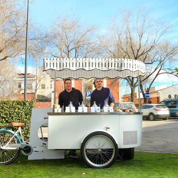Two men stand behind a mobile coffee cart on a sunny day.
