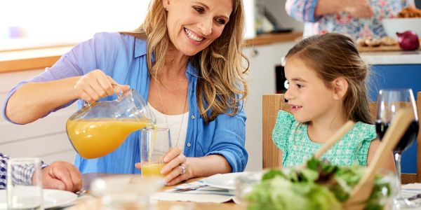 A woman pours juice for a smiling girl at the dining table.