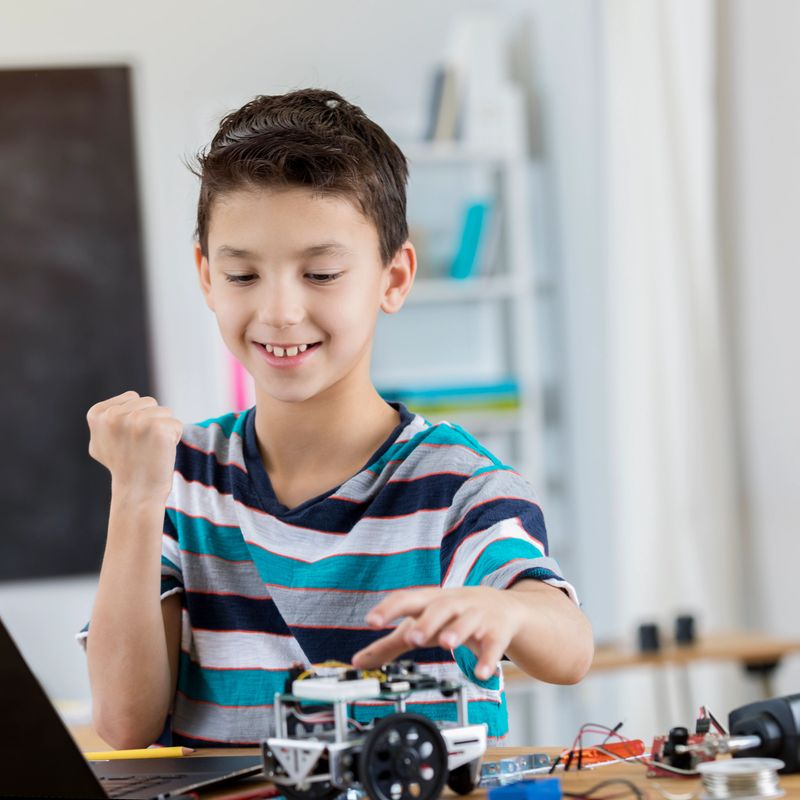 Happy mixed race elementary school student is proud of robot he built in engineering class. He fist pumps while smiling proudly.