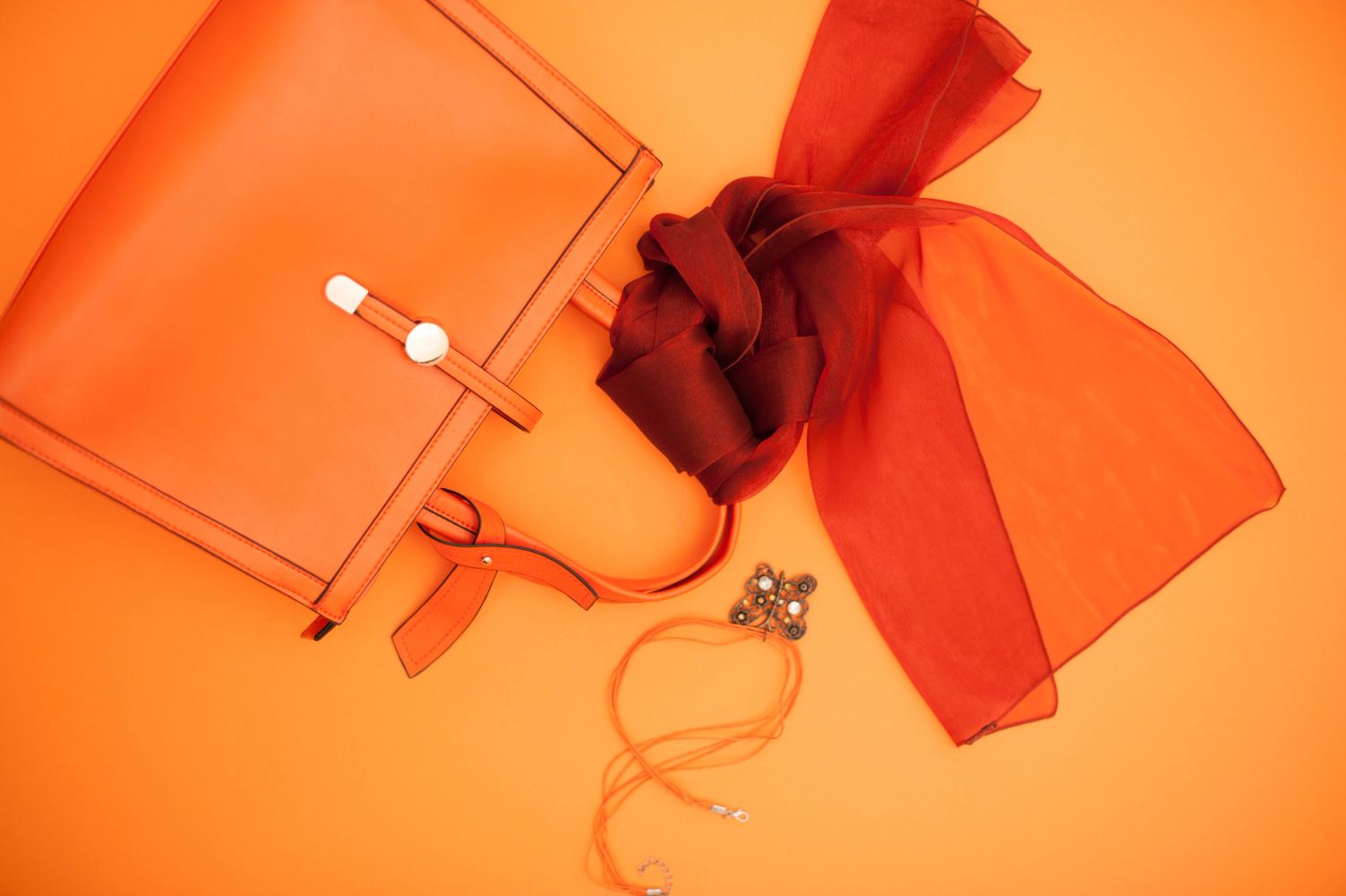 Orange handbag, red scarf, and jewelry on orange background.