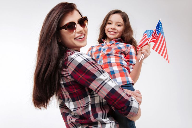 Sharing our joy with you. Young smiling delighted mother holding her daughter and celebrating American national holiday while standing against white background and expressing positivity