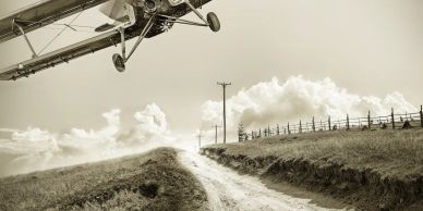 Vintage airplane flying over a dirt road in a countryside setting.