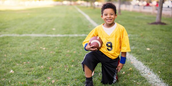 Young boy in yellow jersey kneeling on football field holding a football.