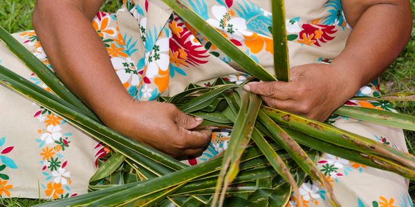 Hands weaving green palm leaves into a pattern over a floral dress.