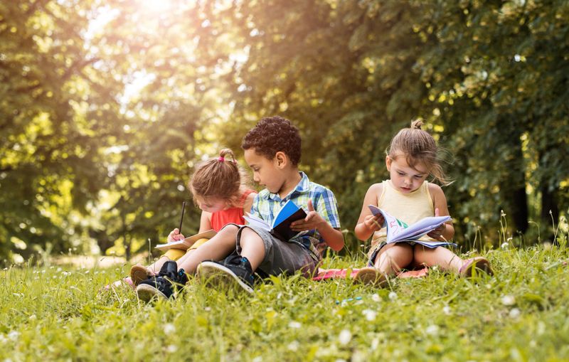 Three small children sitting in grass at the park with their notebooks. Focus is on girl on the right.