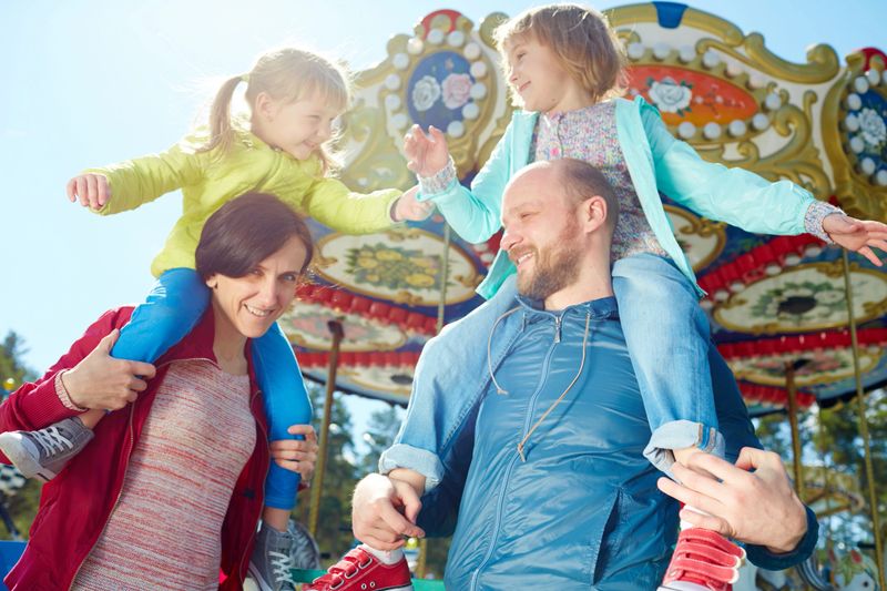 Carefree little girls playing with each other while sitting on shoulders of their smiling middle-aged parents, colorful merry-go-round observed on background