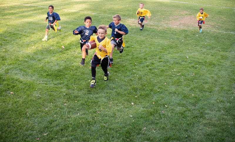 A young child and flag football player runs with the football scoring a touchdown while keeping the other team from pulling his flags. Image taken on an American football field in Utah, USA.