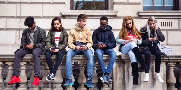 Six teenagers sitting on a ledge, all looking at their smartphones.