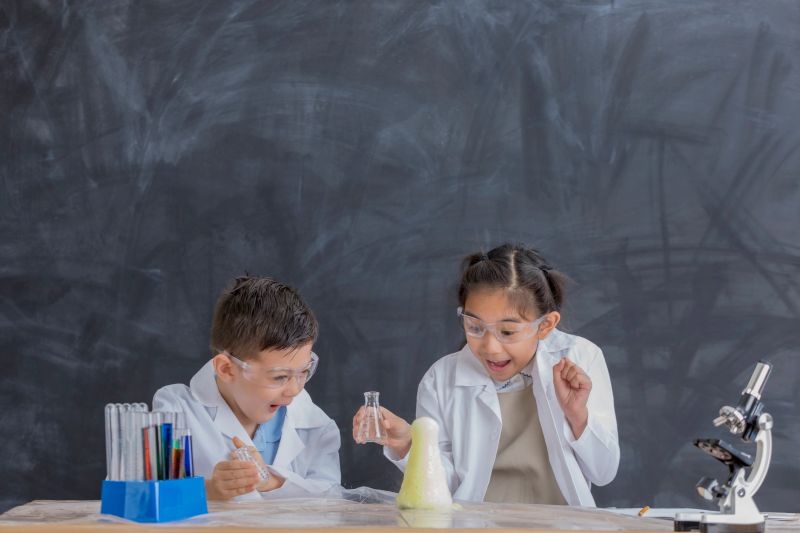 Elementary age Asian brother and sister watch for a chemical reaction while conducting chemistry experiment. They are wearing safety glasses and lab coats.