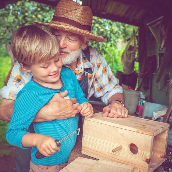 An elderly man helps a young boy build a wooden birdhouse outdoors.