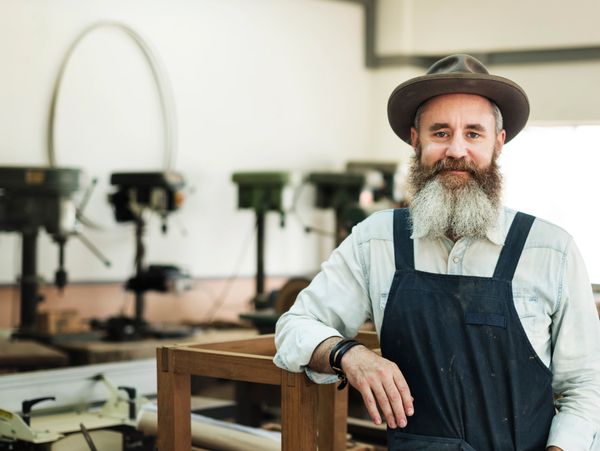 Bearded craftsman in hat and apron in woodworking shop.