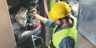 Worker in a yellow hard hat checking a control panel with a tablet in hand.