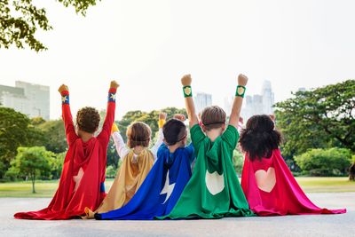 Children dressed as superheroes raise their fists in the air outdoors.