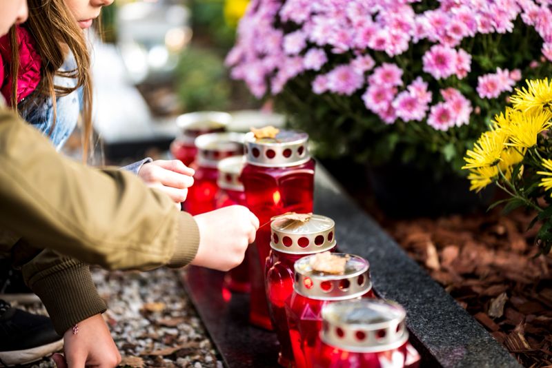 Two kids by the grave. There are a lot of grave candles in a row and one of the kids is holding a leaf on the top of the candle. There are some flowers in the background.