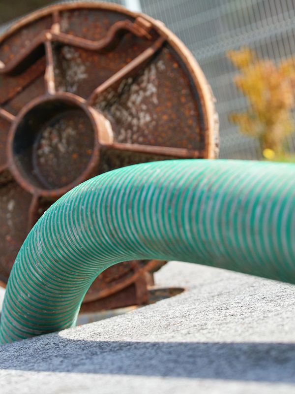 A close-up view of a large green industrial drain hose inserted into an outdoor sewer or clean-out opening. Behind it sits a rusted metal access cover, partially removed to allow service. The scene suggests professional drain cleaning or sewer line maintenance, highlighting heavy-duty equipment used to clear blockages and restore proper flow.