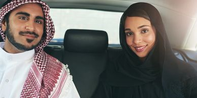 A smiling Middle Eastern couple sitting in a car's backseat, dressed in traditional attire.