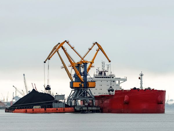 Large red cargo ship docked with cranes unloading coal at a port.