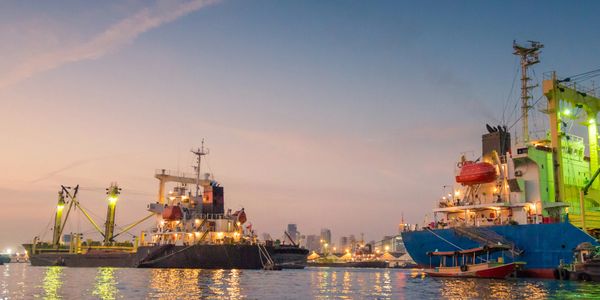 Cargo ships docked at a harbor during sunset with city lights reflecting on the water.