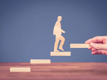 Une main place une marche sous une silhouette en bois pour l’aider à gravir des étapes.