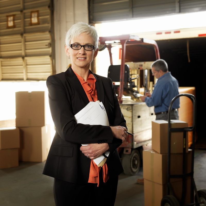 Portrait of businesswoman in a warehouse looking at the camera and holding documents with a colleague working in the background.