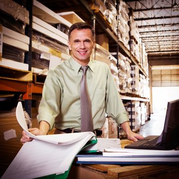 Smiling warehouse manager reviewing documents with a laptop in storage area.