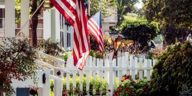 Two American flags adorn a white picket fence in a garden.