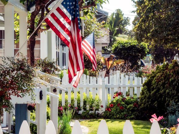 Two American flags hang on white picket fences in a sunny, flower-filled neighborhood.
