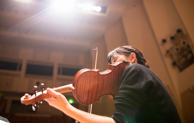 A musician plays the violin under bright stage lights in a concert hall.