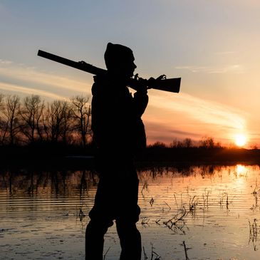 Silhouette of a hunter with a rifle at sunset by a marsh.