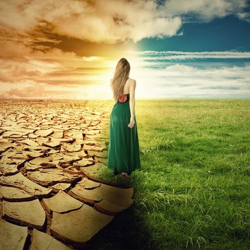 A woman stands between dry cracked earth and lush green grass under a dramatic sky.