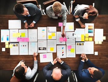 A team collaborates over documents and sticky notes on a wooden table.