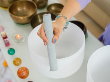 Hand playing a white crystal singing bowl with a mallet on a glass table.