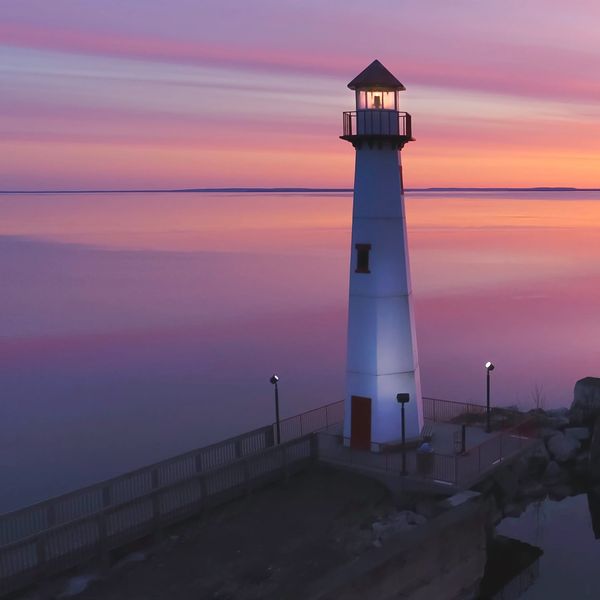 A lighthouse stands tall against a vibrant pink and purple sunset over calm waters.
