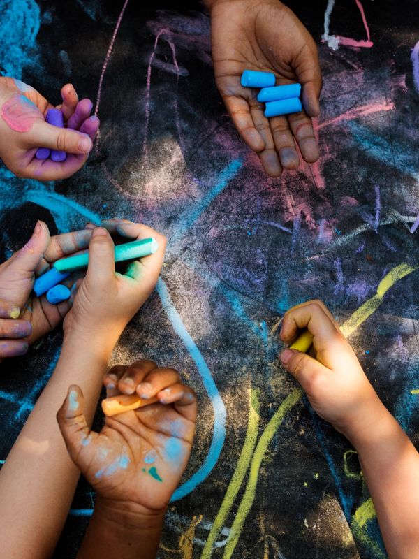 Children's hands drawing colorful chalk art on pavement.