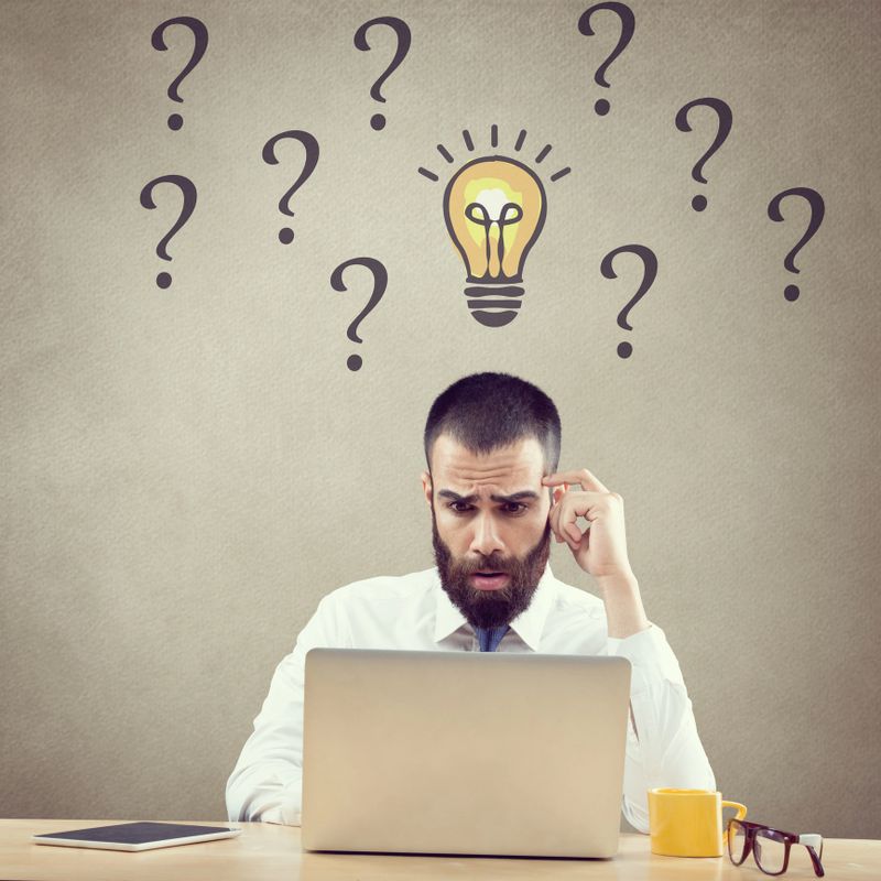 Confused businessman working on laptop in front of light bulb and question marks symbol